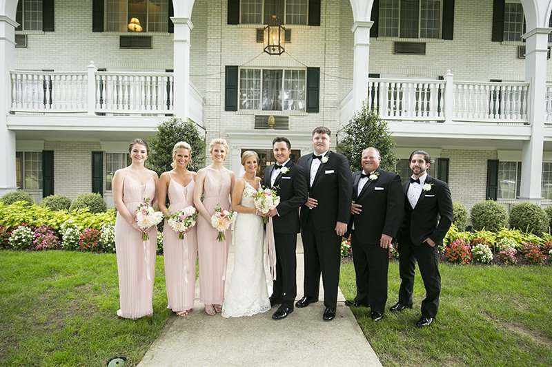 Bridal party posing in front of historic hotel with white bricks, black shutters, colonnade and manicured garden