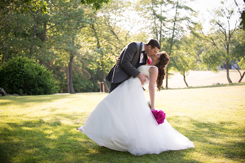 Groom dipping and kissing bride on grassy field, shaded by a tree