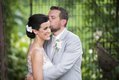 Groom tenderly kissing bride's forehead, posing outdoors with greenery behind