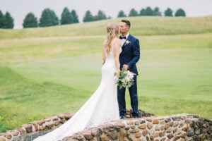 Wedding day photo of a bride and groom kissing on a rustic stone bridge over a small creek in the middle of a green field.